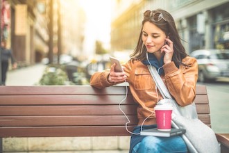 Image of a smiling woman sitting on a bench with headphones looking at a phone.