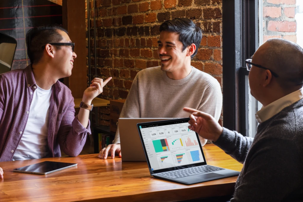 Three coworkers sitting at a table talking over a Microsoft Surface laptop.