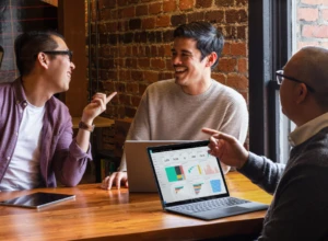 Three coworkers sitting at a table talking over a Microsoft Surface laptop.