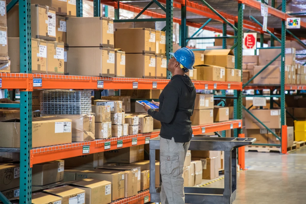Male worker wearing hardhat using tablet and looking at boxes in warehouse.