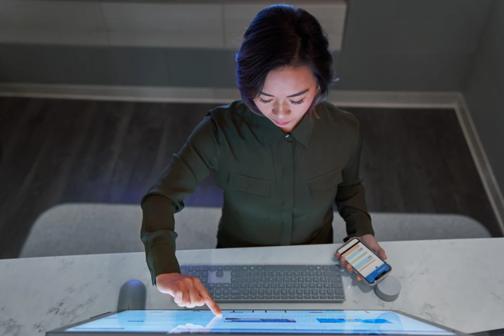 Top down view of a woman wearing a dark shirt in a dim office scrolling or working on a Microsoft Surface Studio and holding a phone. Keywords: touch screen, desktop, threat protection, secure score, monitoring, Microsoft Security collection