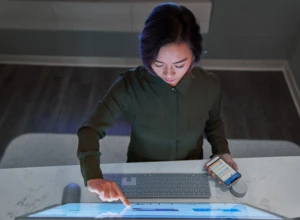 Top down view of a woman wearing a dark shirt in a dim office scrolling or working on a Microsoft Surface Studio and holding a phone. Keywords: touch screen, desktop, threat protection, secure score, monitoring, Microsoft Security collection