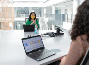 Two female employees wearing face masks and talking in a conference room while social distancing. Hybrid Workplace collection. Keywords: Hybrid Workplace, Hybrid Worksite, Return to Workplace, Return to Work, Return to Worksite, Return to Office, Return to Campus, Reopening, Back to work, COVID-19, COVID-19 policies, COVID, Coronavirus, Pandemic, Health, Health and Safety, Face Covering, Face Mask, Masks, Masks at Work, Masks in Workplace, Open Workspace, Open Workspace Policies, Social Distancing, Social Distancing Policies, Social Distance, Physical Distancing.