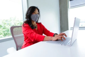 A female employee wearing a face mask and working on her Surface Book laptop at her desk. Hybrid Workplace collection.
