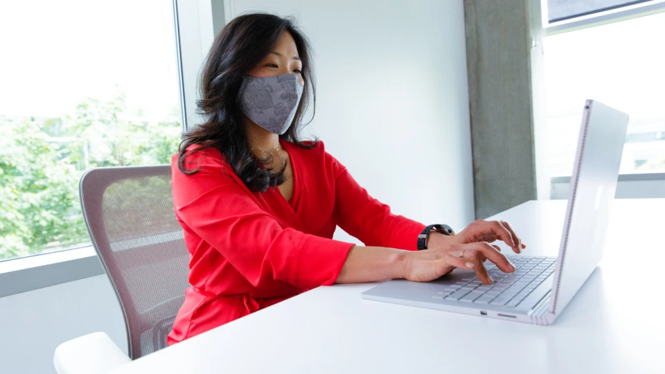 A female employee wearing a face mask and working on her Surface Book laptop at her desk. Hybrid Workplace collection.