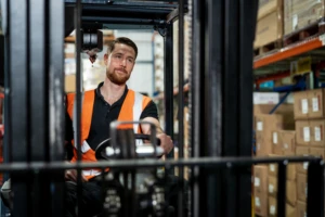 Man wearing an orange vest in a manufacturing or warehouse driving a forklift. Key words: tele conferencing, cloud calling, Microsoft Teams, meeting, conference call, phone system, video system, Business Voice, Manufacturing, Firstline Worker, Delivery, Tracking