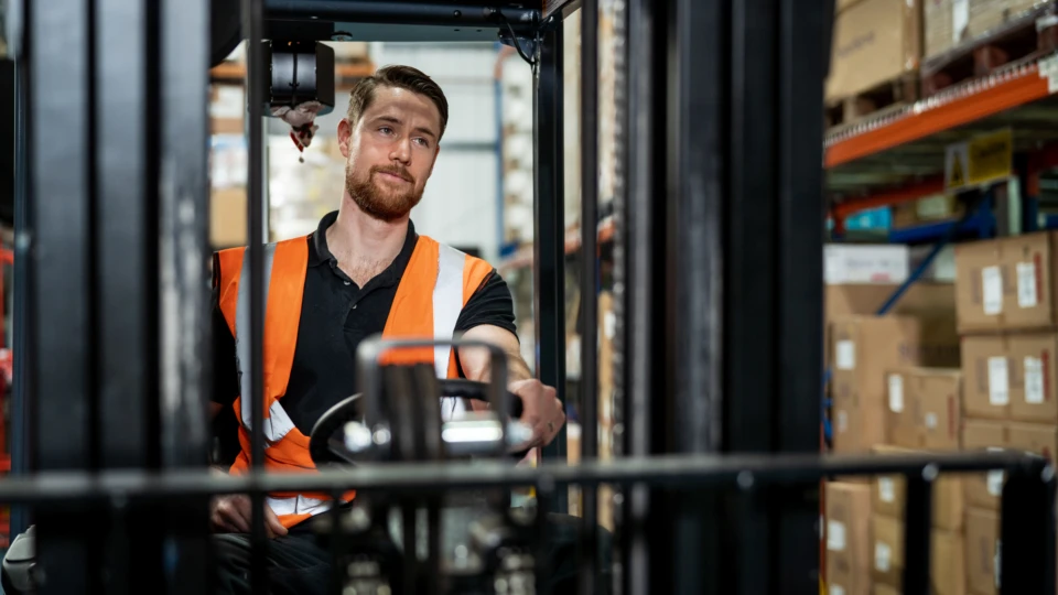 Man wearing an orange vest in a manufacturing or warehouse driving a forklift. Key words: tele conferencing, cloud calling, Microsoft Teams, meeting, conference call, phone system, video system, Business Voice, Manufacturing, Firstline Worker, Delivery, Tracking