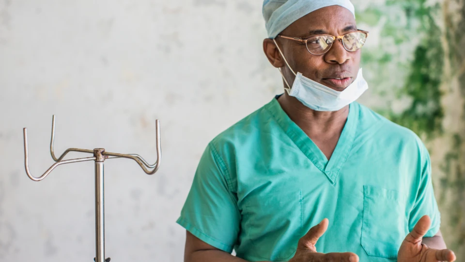 Real people. Male doctor wearing scrubs, glasses, and face mask standing and speaking in medical facility in Liberia.