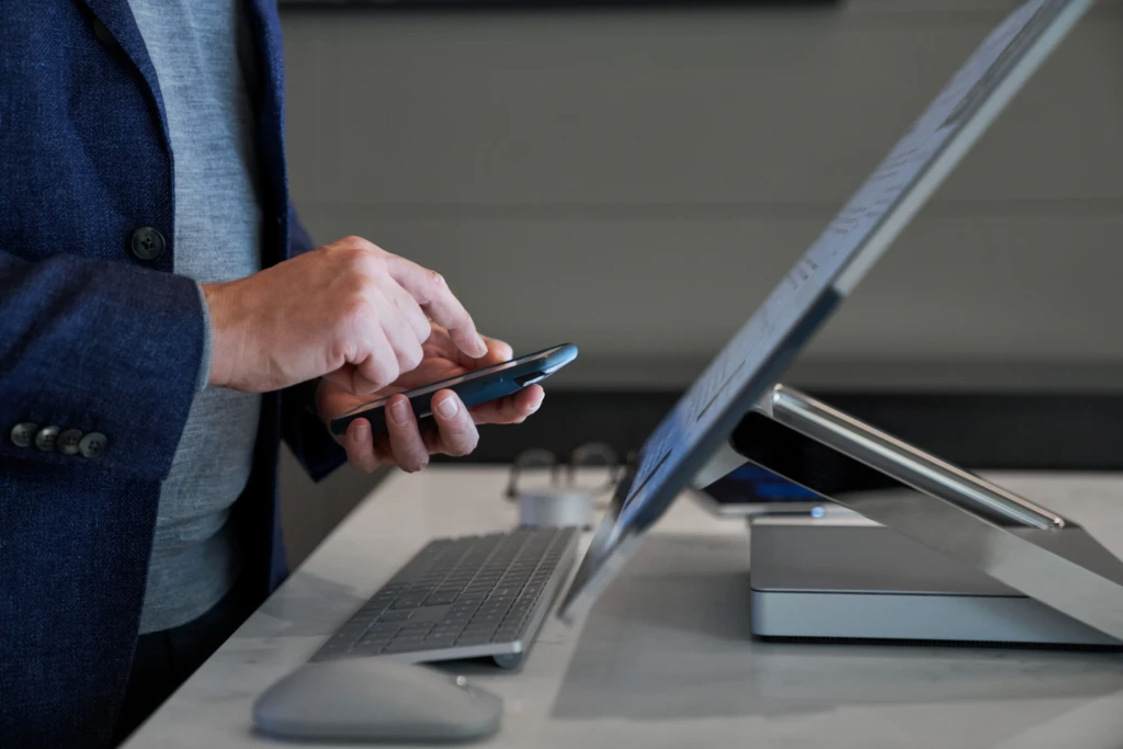 Side view close-up of a man typing on his phone while standing behind a Microsoft Surface Studio. Keywords: touch screen, desktop, multi-device, mouse, keyboard, hands, cross platform, MFA, multi-factor authentication, threat protection, secure score, monitoring, Microsoft Security collection
