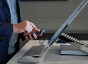 Side view close-up of a man typing on his phone while standing behind a Microsoft Surface Studio. Keywords: touch screen, desktop, multi-device, mouse, keyboard, hands, cross platform, MFA, multi-factor authentication, threat protection, secure score, monitoring, Microsoft Security collection