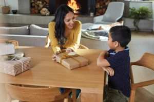 Mother and son wrap gifts at table in living room with fireplace.
