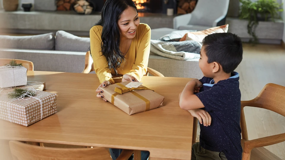 Mother and son wrap gifts at table in living room with fireplace.