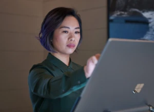 Side profile of a woman wearing a dark shirt in a dim office reaching up and working on a Microsoft Surface Studio. Keywords: touch screen, desktop, cloud security, threat protection, secure score, monitoring, Microsoft Security collection