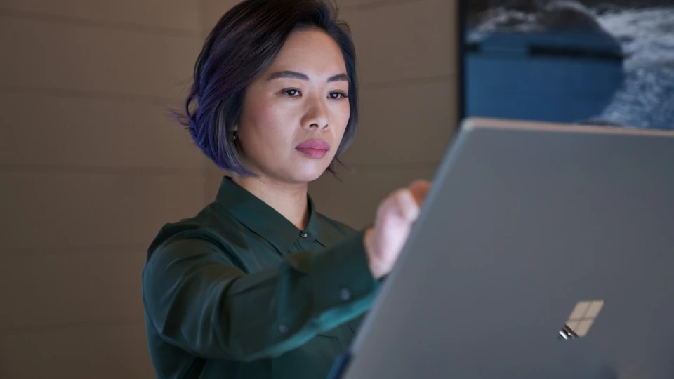 Side profile of a woman wearing a dark shirt in a dim office reaching up and working on a Microsoft Surface Studio. Keywords: touch screen, desktop, cloud security, threat protection, secure score, monitoring, Microsoft Security collection