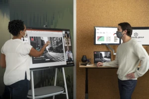 Two adults in masks collaborating on a PowerPoint presentation in an office while using a Microsoft Surface Hub 2S 50” device during a Teams video call.