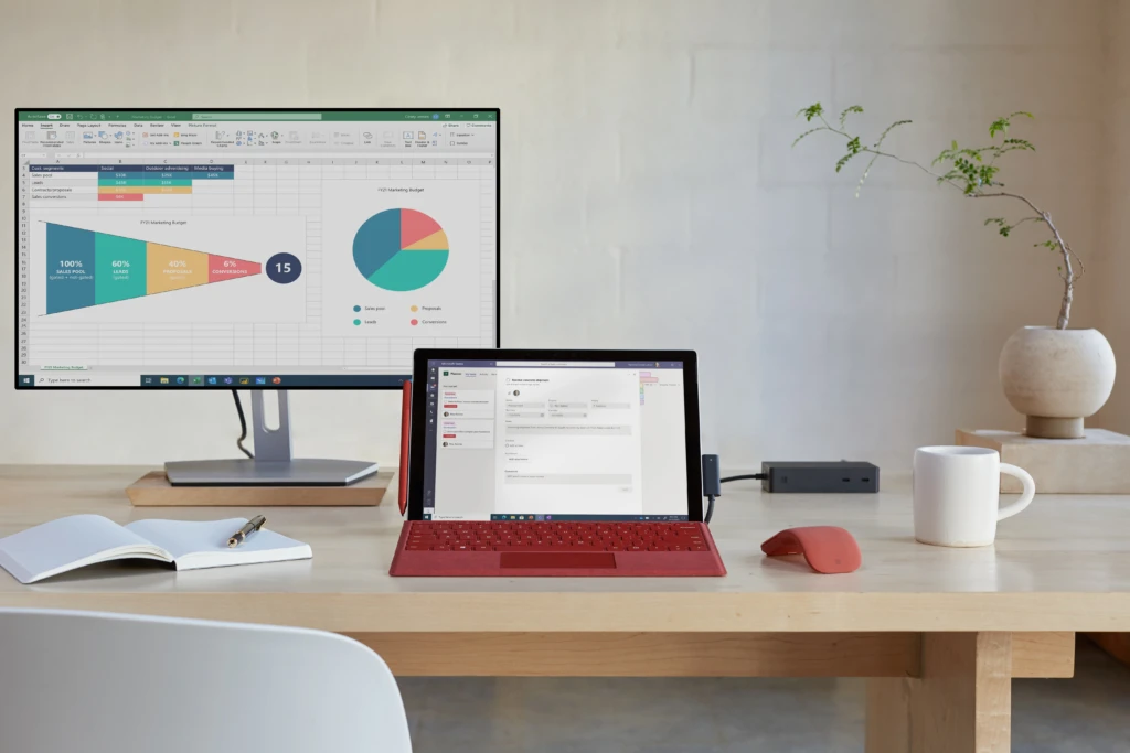 Front view of a Surface Pro 7+ with a poppy red keyboard and poppy red Surface Arc Mouse. Surface Pro 7+ screen is displaying Microsoft Teams and the monitor is displaying Microsoft Excel.
