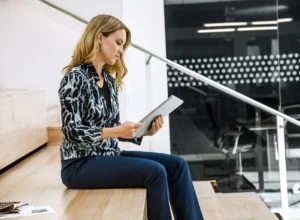 Woman sitting on steps in open office building. She is using a tablet.
