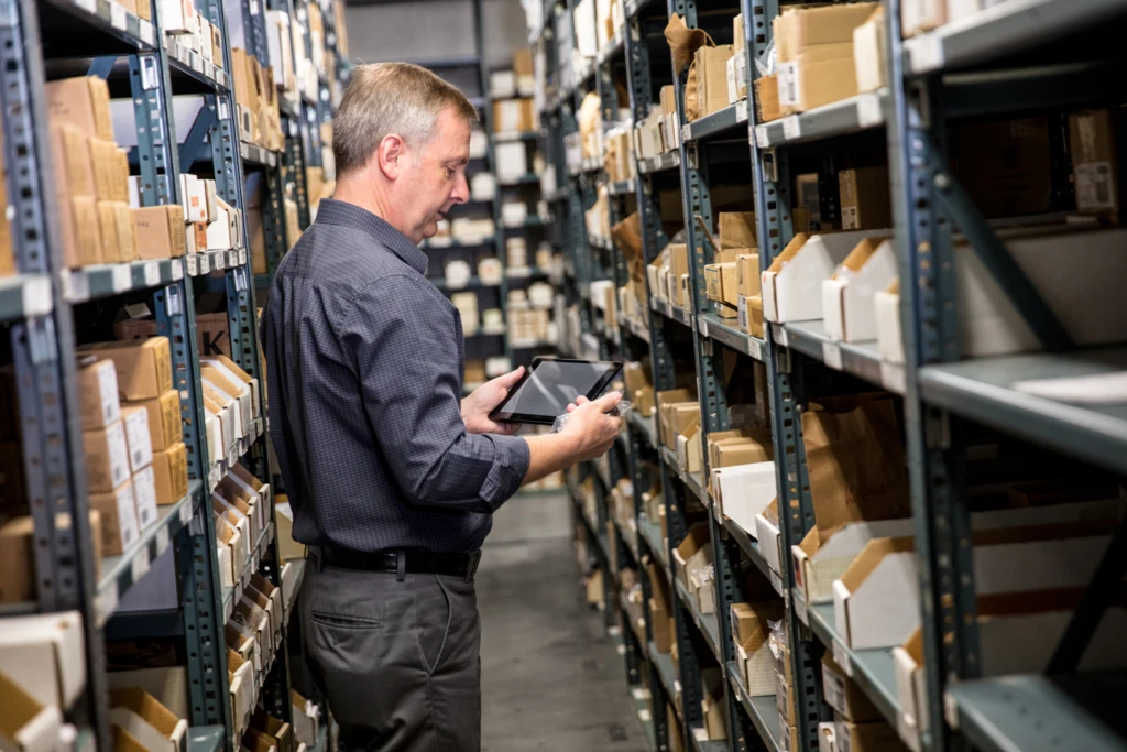 Male employee in warehouse storage room holding a tablet in one hand and small item in the other. He is standing between metal warehouse racks filled with pacakges.