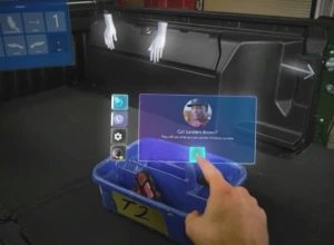 A user’s mixed reality view from their HoloLens, showing their hand using touch controls for starting a holographic video call with a remote colleague.
