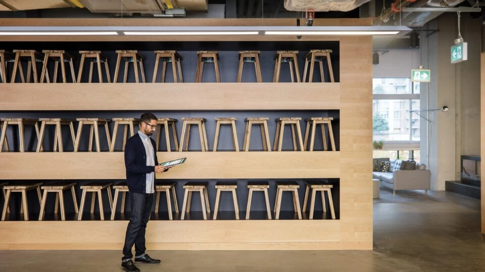 Male small business worker standing in front of three large shelves filled with dozens of wood stools. He is using an HP convertible laptop (opened as tablet) and pen. Screen is partially shown depicting bar charts and graphs.