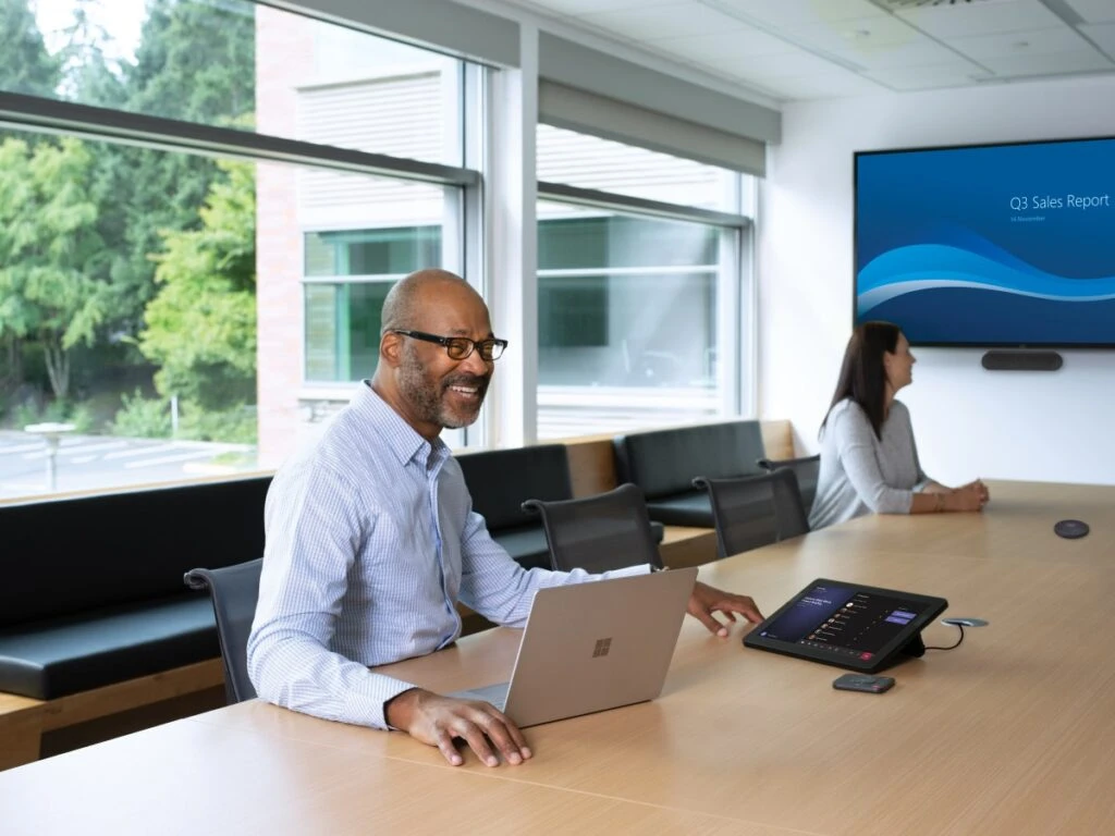 One male and one female in a large conference room featuring an HP Teams Meeting Rooms touch display joined to a Teams Meeting. Screensharing on a large mounted display and one Surface Devices in view.