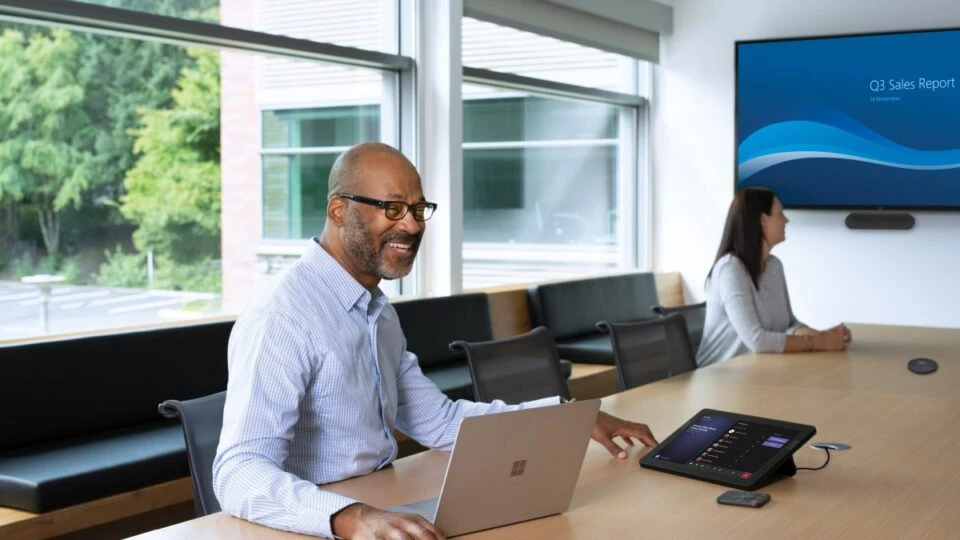 One male and one female in a large conference room featuring an HP Teams Meeting Rooms touch display joined to a Teams Meeting. Screensharing on a large mounted display and one Surface Devices in view.