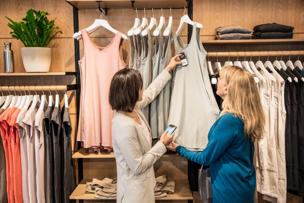 Photo of a female store employee using phone standing next to female consumer in front of retail clothing display. The employee is checking the price tag.