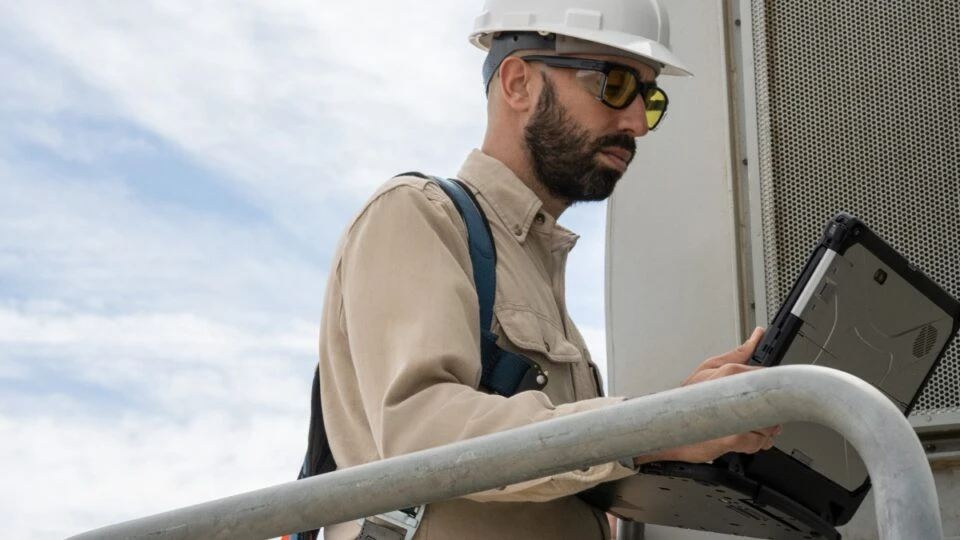 Field engineer viewing data on a laptop after inspecting turbines on a wind farm. Keywords: Dynamics 365; operations; green energy; outside; outdoors; on the go; remote assist; training; man; wearing hard hat and safety gear