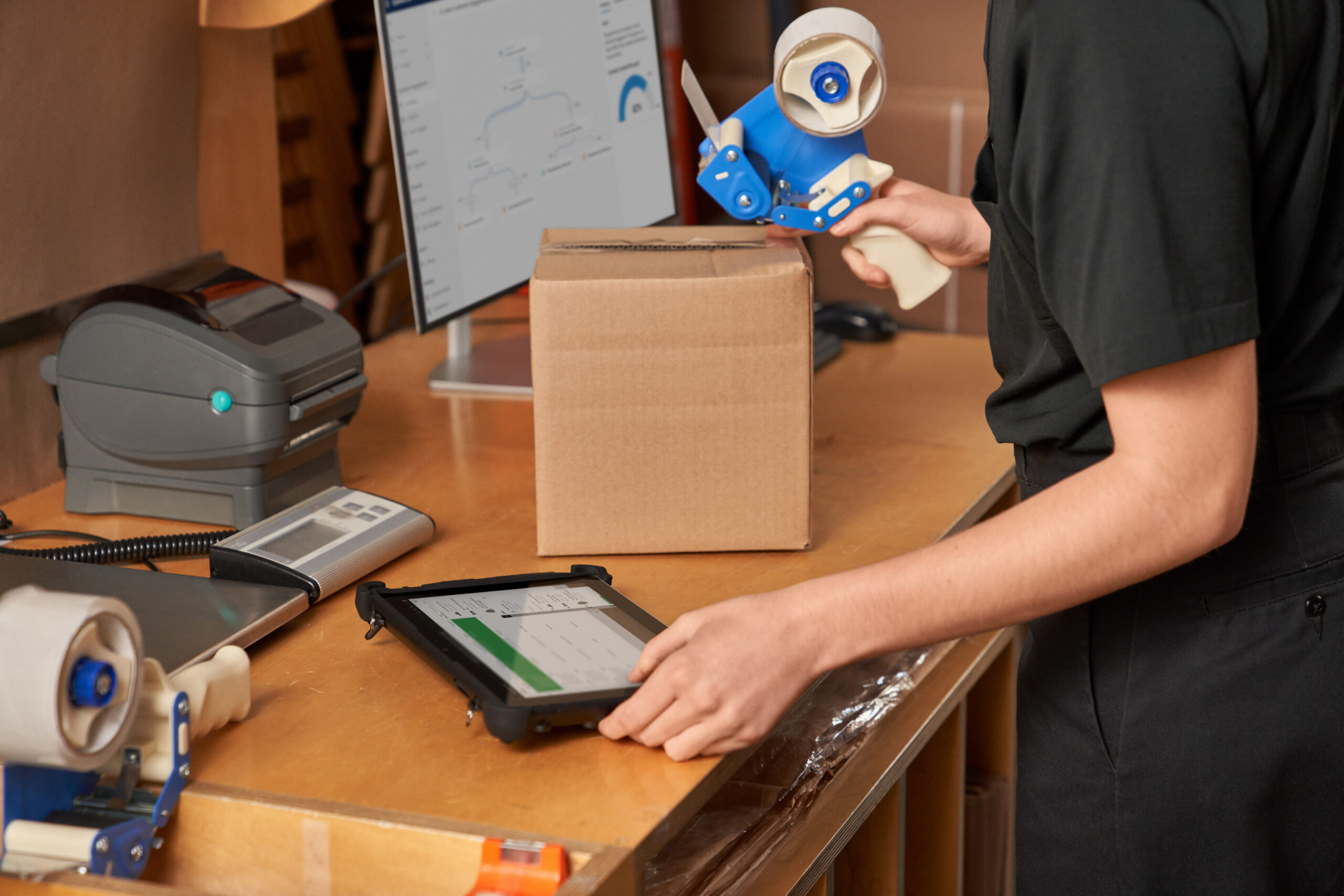 Customer service rep fulfilling, packaging and shipping online orders in the storeroom with a finance and operations mobile app on a phone and a desktop display.