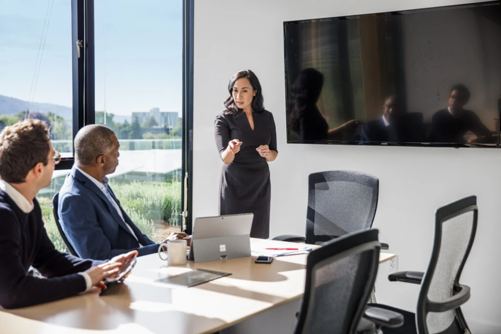 Female corporate executive giving speech to two male coworkers at conference room table in corporate office.