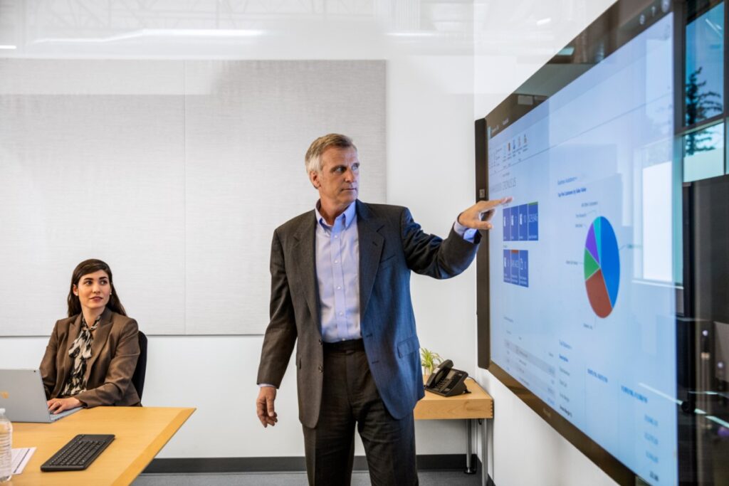 Male businessman in suit giving presentation in office conference room. He is pointing at a large monitor screen, which displays a pie chart and several statistics/monetary values.