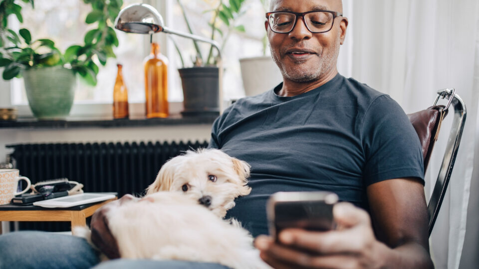 Photo of a senior male using his Apple Messages app while sitting with dog in room at home.