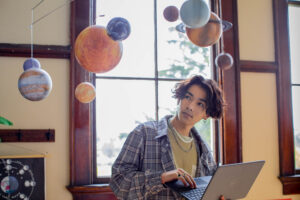 Photo of a student in a classroom working on an open laptop and looking at a solar system mobile hanging from the ceiling.