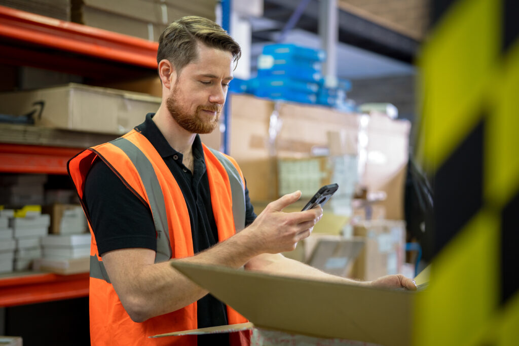 Photo of a man wearing an orange vest in a warehouse who is doing inventory on his mobile phone.