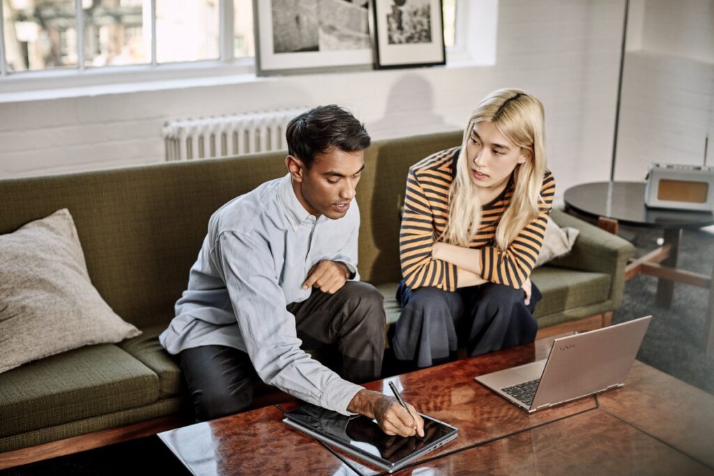 A man and women sitting on a couch in an office setting, collaborating on a project with their Lenovo Yoga; one set up as a Laptop and the other being used as a tablet.
