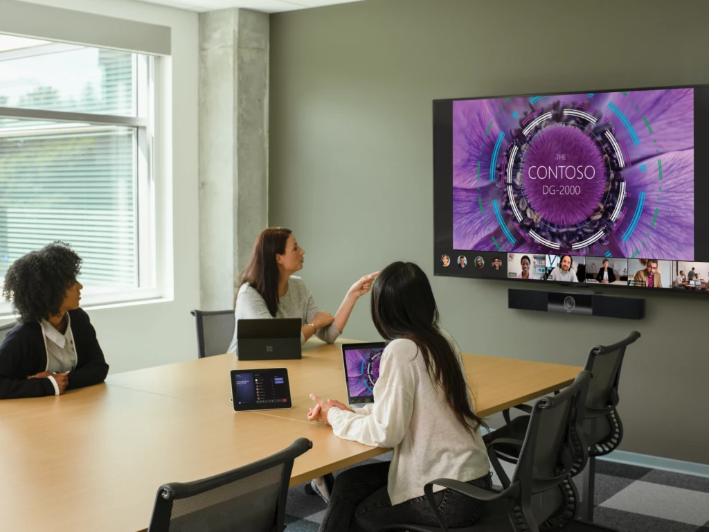 Three females in a large conference room featuring an Poly Teams Meeting Rooms touch display joined to a Teams Meeting. Screensharing and remote participants shown on a large mounted display and two Surface Devices in view.