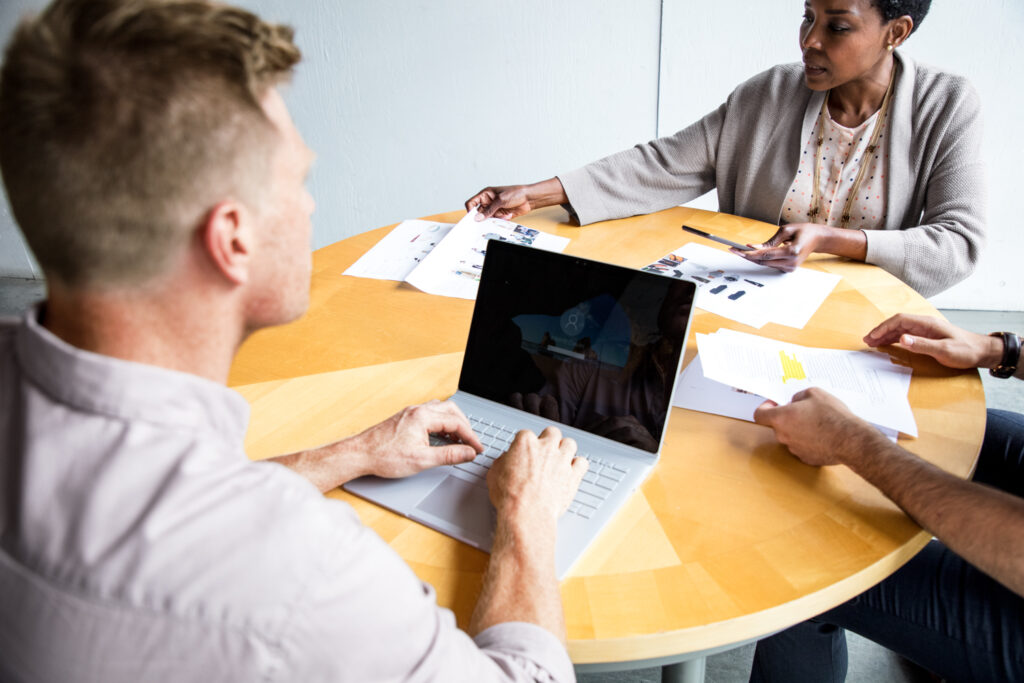 Female and male workers sitting at round table in discussion. A third person's (male) hands are also visible. The man is using a Surface Laptop, while the woman is reviewing paperwork.