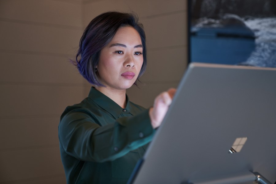 Side profile of a woman wearing a dark shirt in a dim office reaching up and working on a Microsoft Surface Studio.