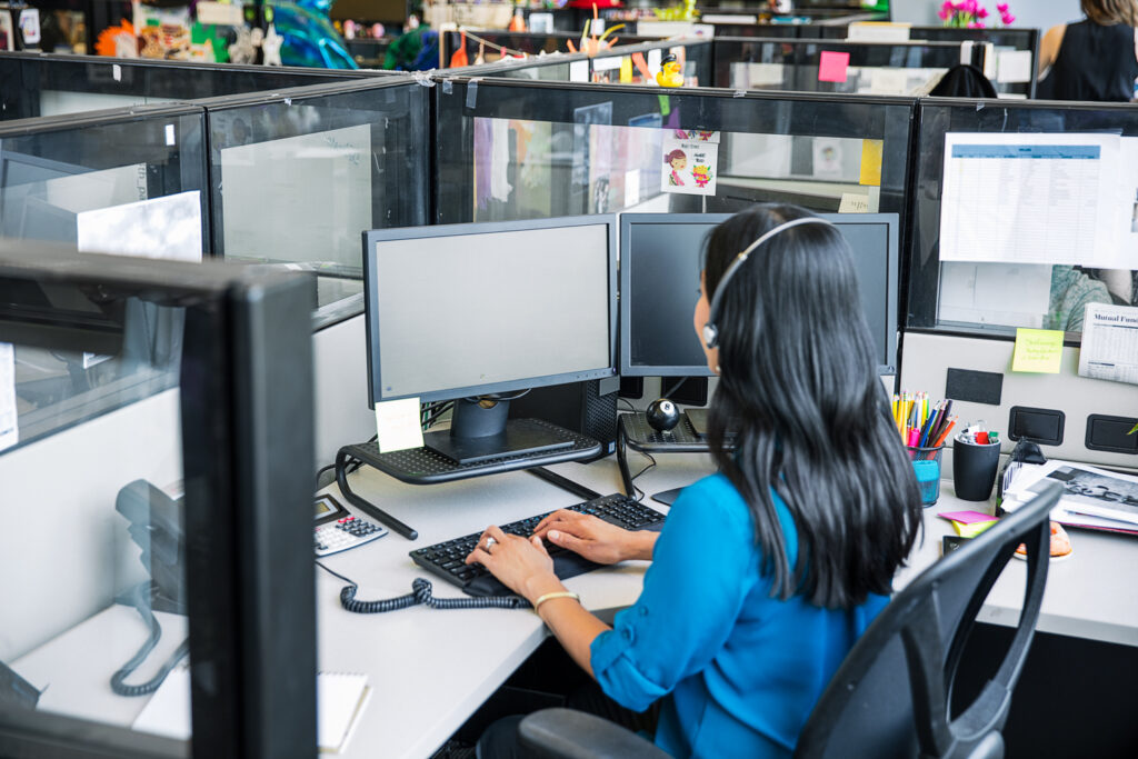 Female call center employee sitting at cubicle desk, using desktop computer with both hands on keyboard. Two monitors shown (no displays shown on screens).