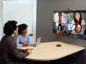 One male and one female in small conference room with a Yealink Microsoft Teams Room on Android device being used for remote video meeting with Teams Meetings. Large mounted display showing remote participants and one Surface Device in view.