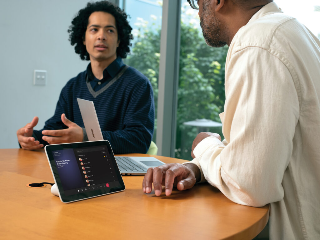 Two males in small conference room with a Poly Microsoft Teams Rooms on Android touch display in a Teams Meeting and one Surface laptop in view.