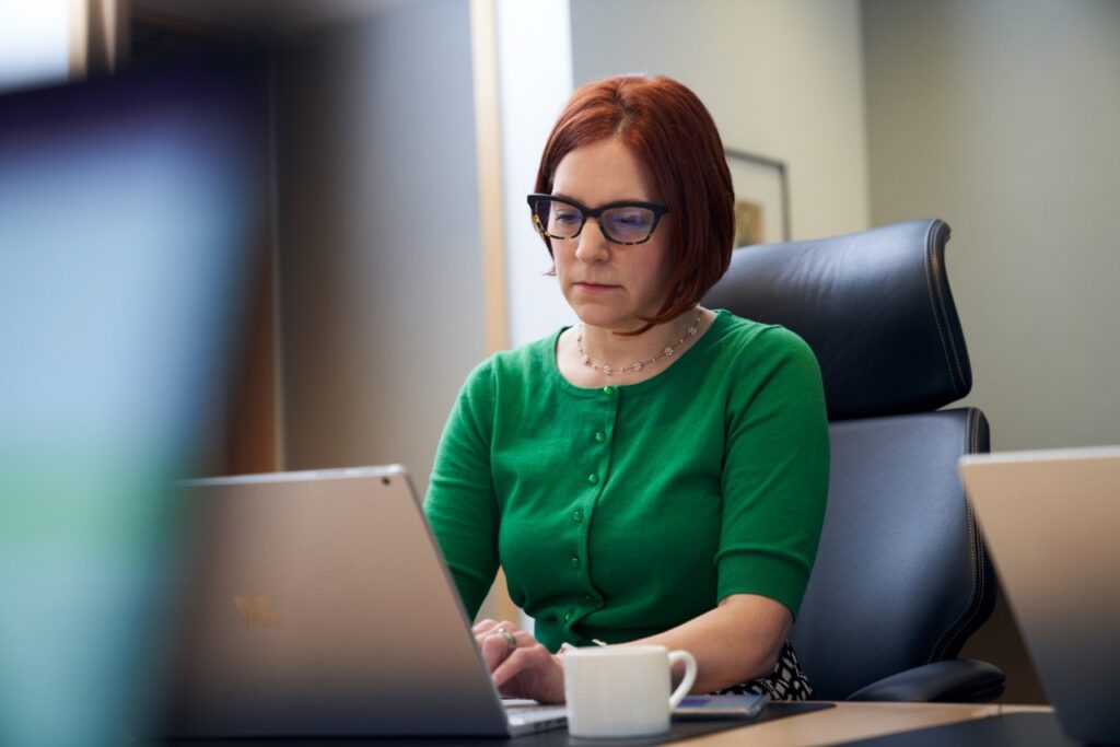 A red-haired woman with a green shirt and glasses, sitting inside an office working on her laptop.