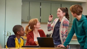 Drei Schüler sind mit einer Lehrerin an einem Laptop. Eine Schülerin erklärt den anderen etwas.
