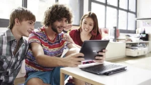drei Studenten, zwei Männer und eine Frau, sitzen an einem Tisch und schauen gemeinsam auf ein Tablet PC