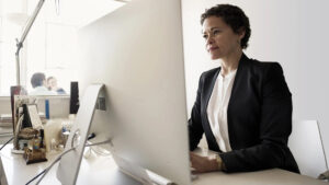 Eine Frau im schwarzen Anzug sitzt vor einem PC am Schreibtisch