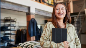 Eine Frau lächelt in einem Klamottenladen und hält einen Tablet.