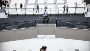 Aerial view of office building‘s interior foyer. Two men are walking across elevated walkway. Man and woman on escalator, various others walking on ground floor below.