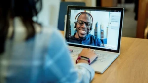 Photograph of a person at a desk using a Surface laptop to make a Microsoft Teams video call with a man who is smiling and wearing a headset.