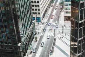 Downtown city street view from high angle inside office building. Cars flow through intersection down one way street. Some pedestrians visible on sidewalks and at crosswalk.