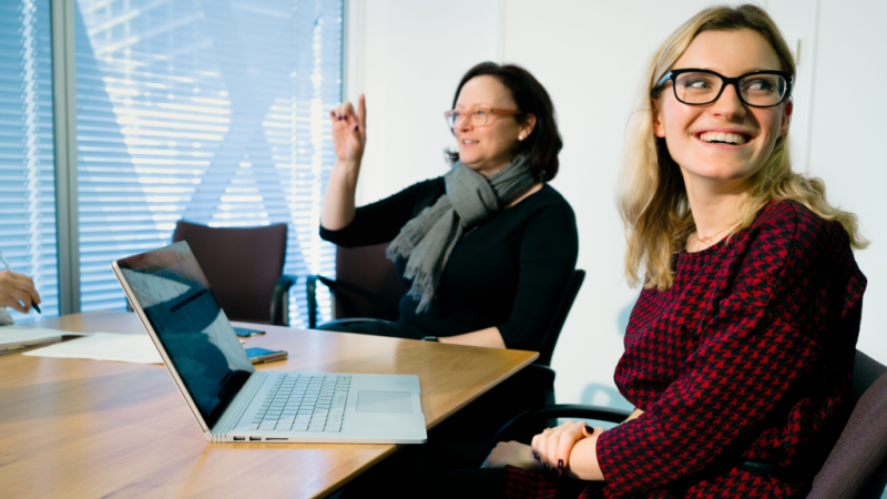 Female office worker with glasses sitting at conference room table, looking over shoulder and smiling. An open Surface Book sits on the table in front of her (screen partially visible but blurry). A female coworker sits behind her, in midst of conversation.